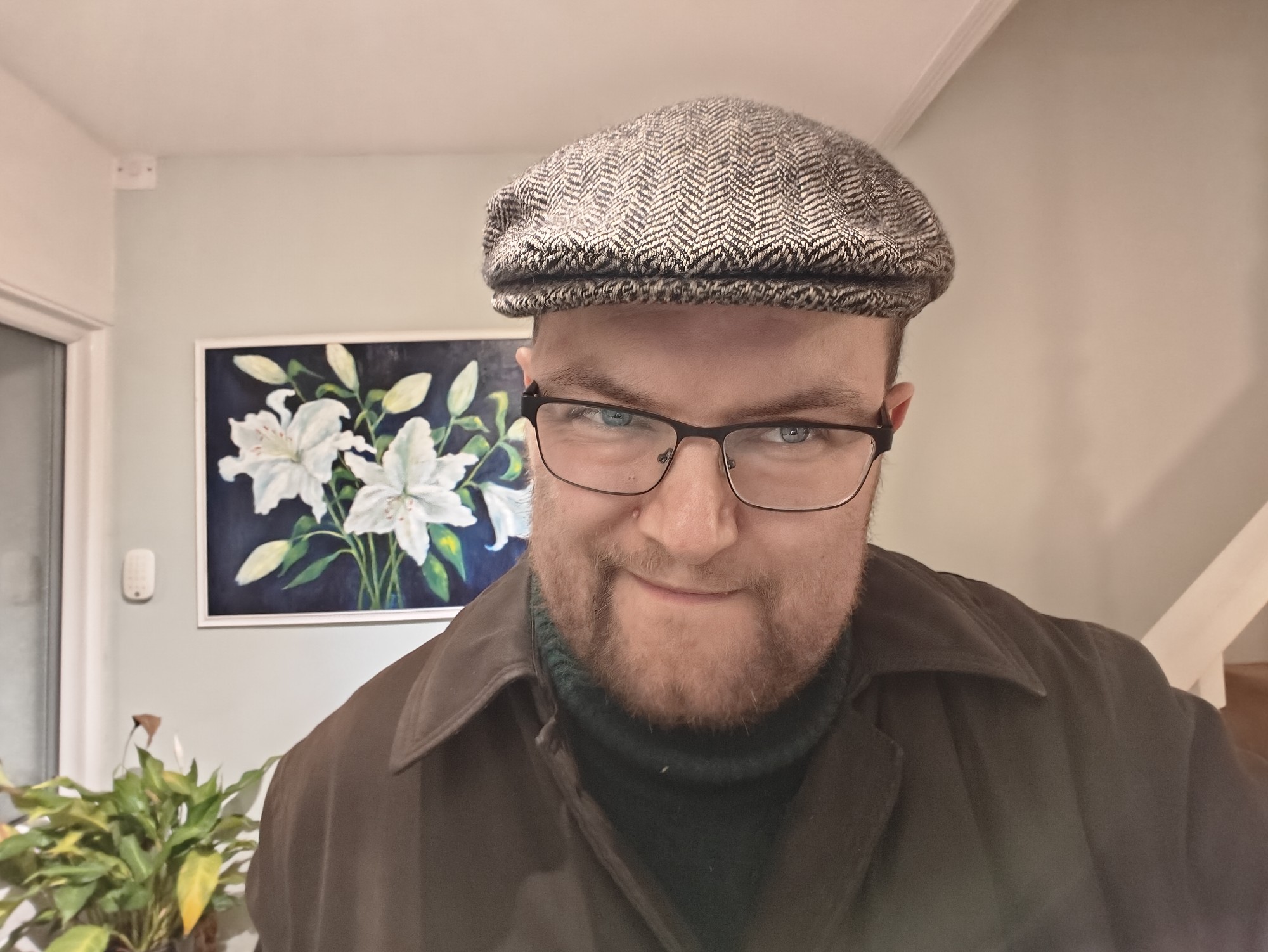 A picture of me, Martin Paul Eve, wearing a coat and flat cap. Behind on the wall is a painted picture of some lillies. 