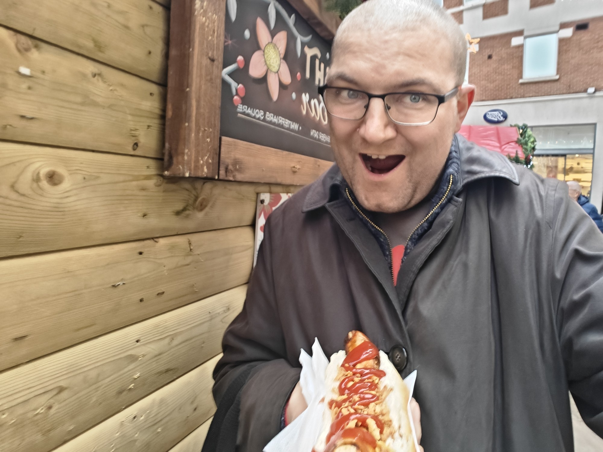 A picture of me, looking happy, wearing glasses, bald head, eating/holding a bratwurst hot dog. 