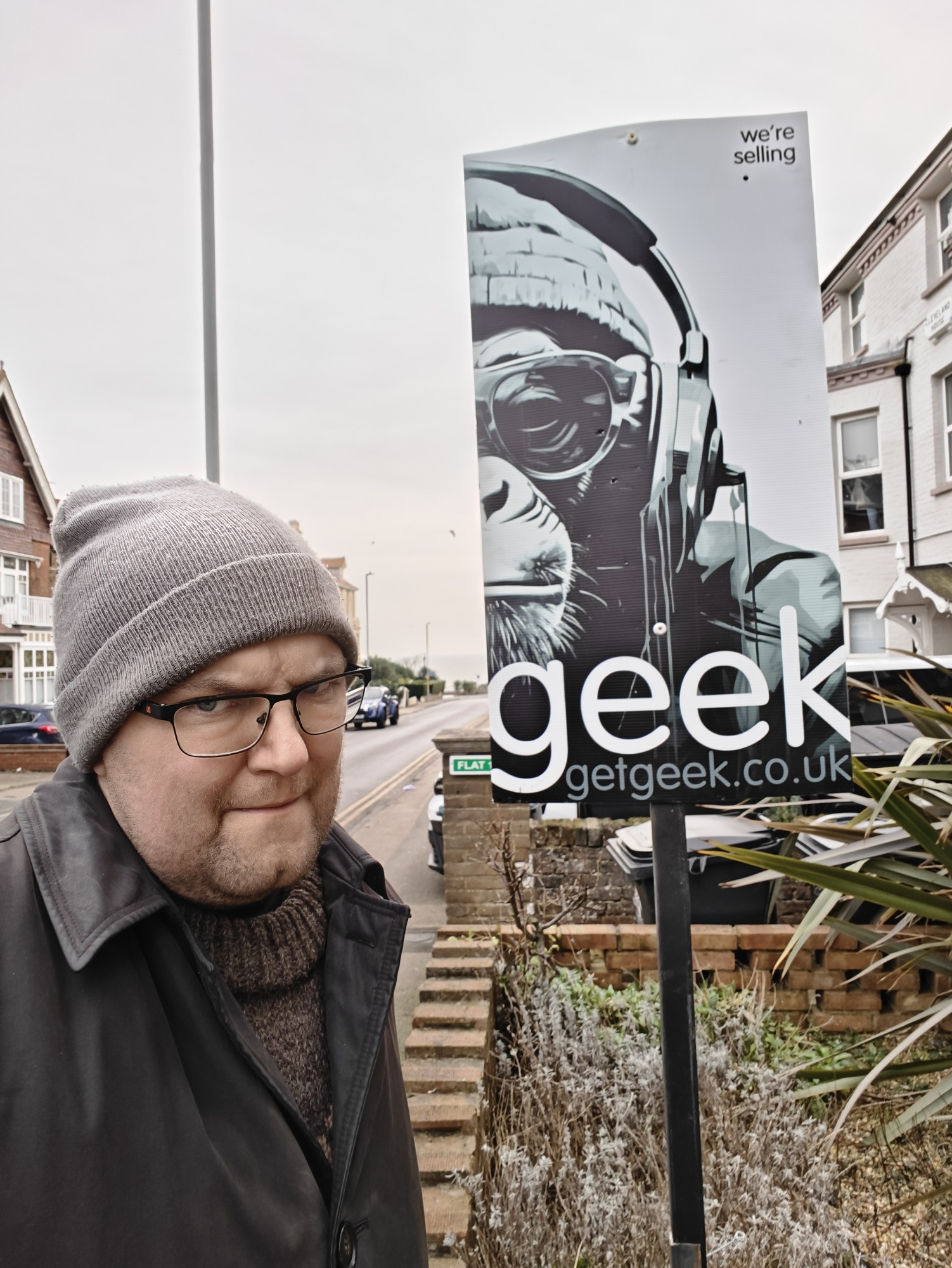 A photograph of me wearing a woolly hat next to an estate agent sign that says "geek". 