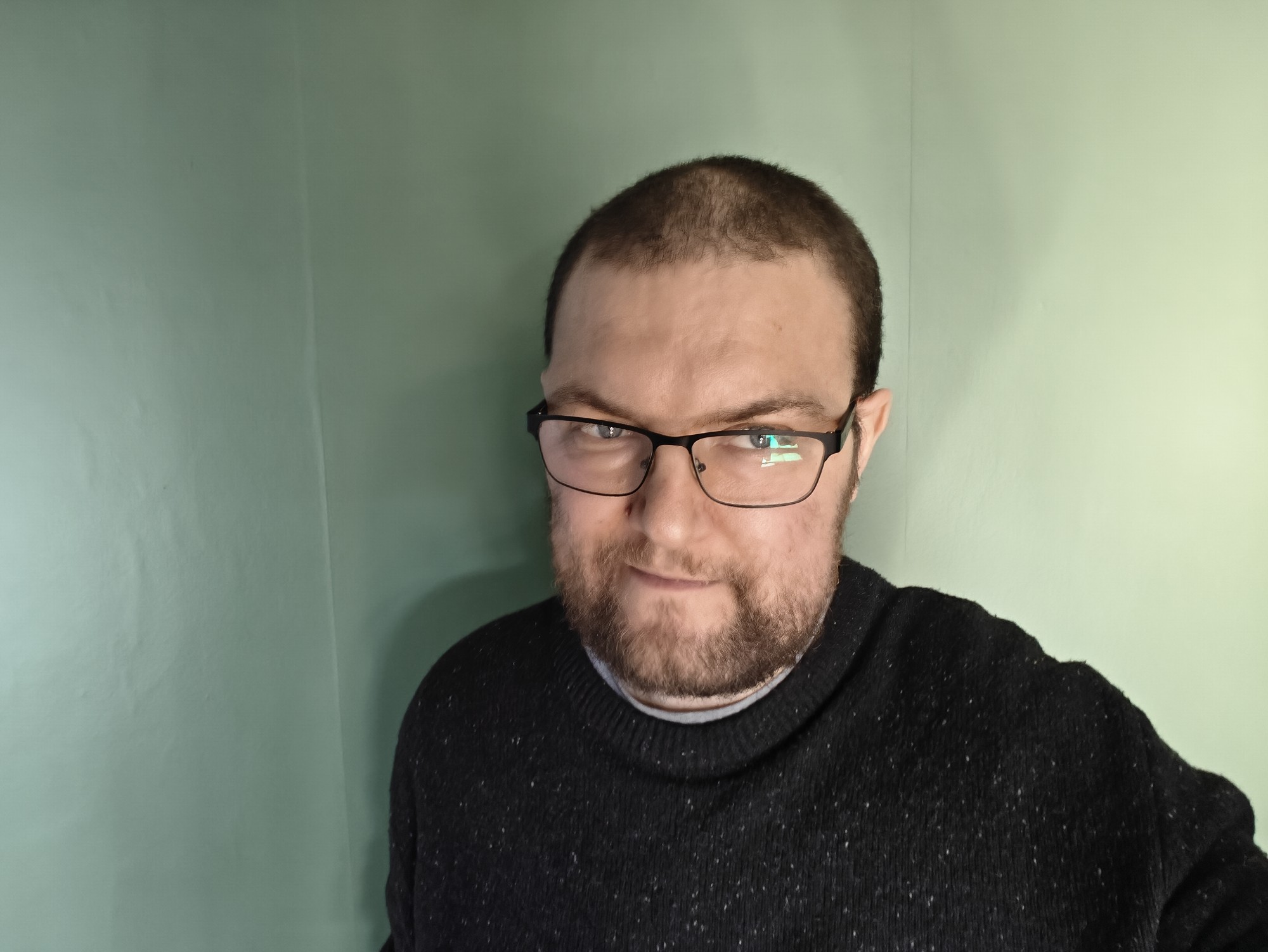 Me - a 40 year old man - wearing glasses with a black jumper against a green background. I have a beard and some short hair with a bald patch. 