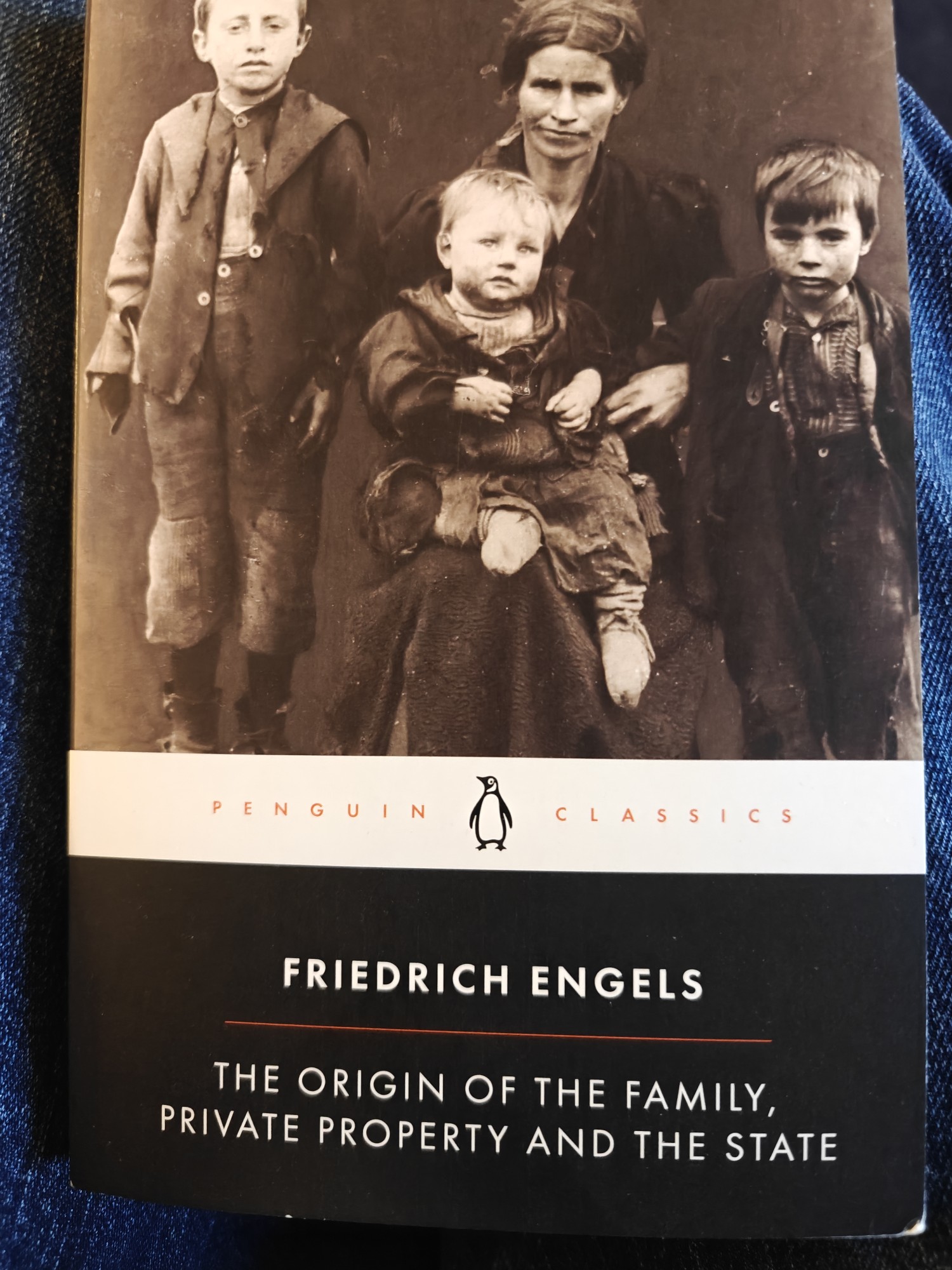 The cover of Friedrich Engels, The Origin of the Family, Private Property and the State. It shows a sepia photograph of three children and a woman, with one child sitting on her knee. The photograph is called "A Starving Family" taken by an unknown photographer around the turn of the 20th century. All the photographic subjects appear grubby and look miserable. 