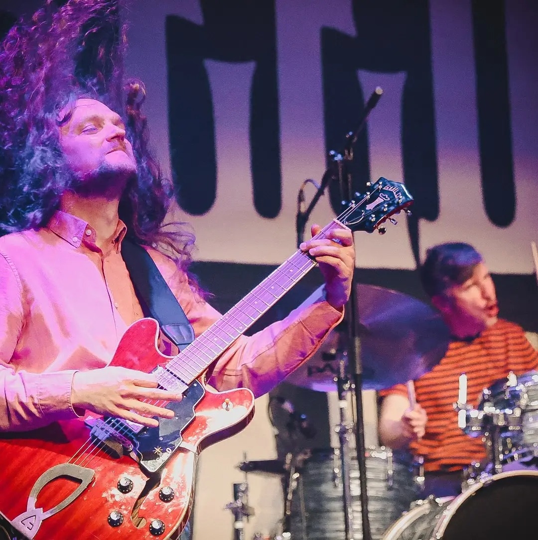 A picture of Rich Hinks playing bass guitar. He has long hair and white skin and a beard. He is wearing a pink shirt. 