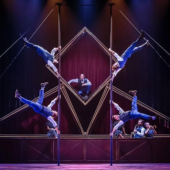 A picture of Cirque Eloize performing, with 5 acrobats climbing poles and inverting their bodies in various ways