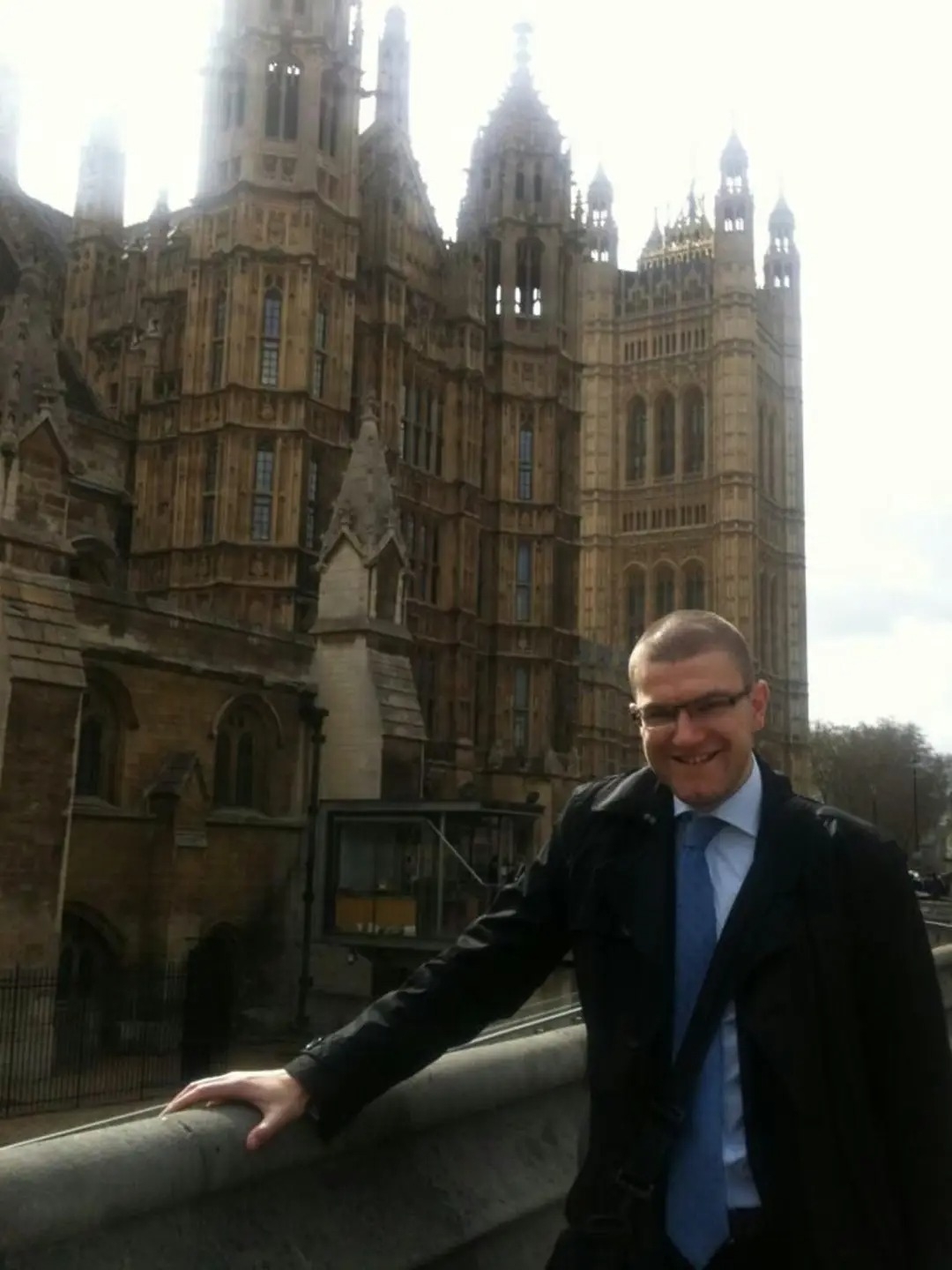 A picture of me outside the House of Commons. I am wearing a suit and blue tie and smiling. The HoC looks like a fantasy castle. 