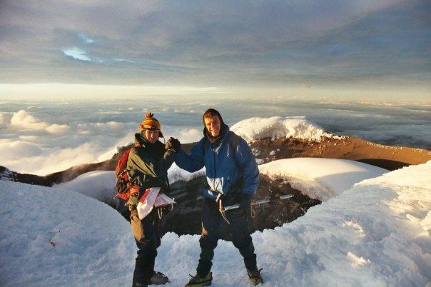 Two men standing on an icy summit with a large volcanic crater behind them. They are holding ice axes. They are holding each other's hands in a locked formation in celebration of reaching the top. I (Martin Eve) am on the right, smiling triumphantly. The other man, Julian Cottee, is wearing a bobble hat.