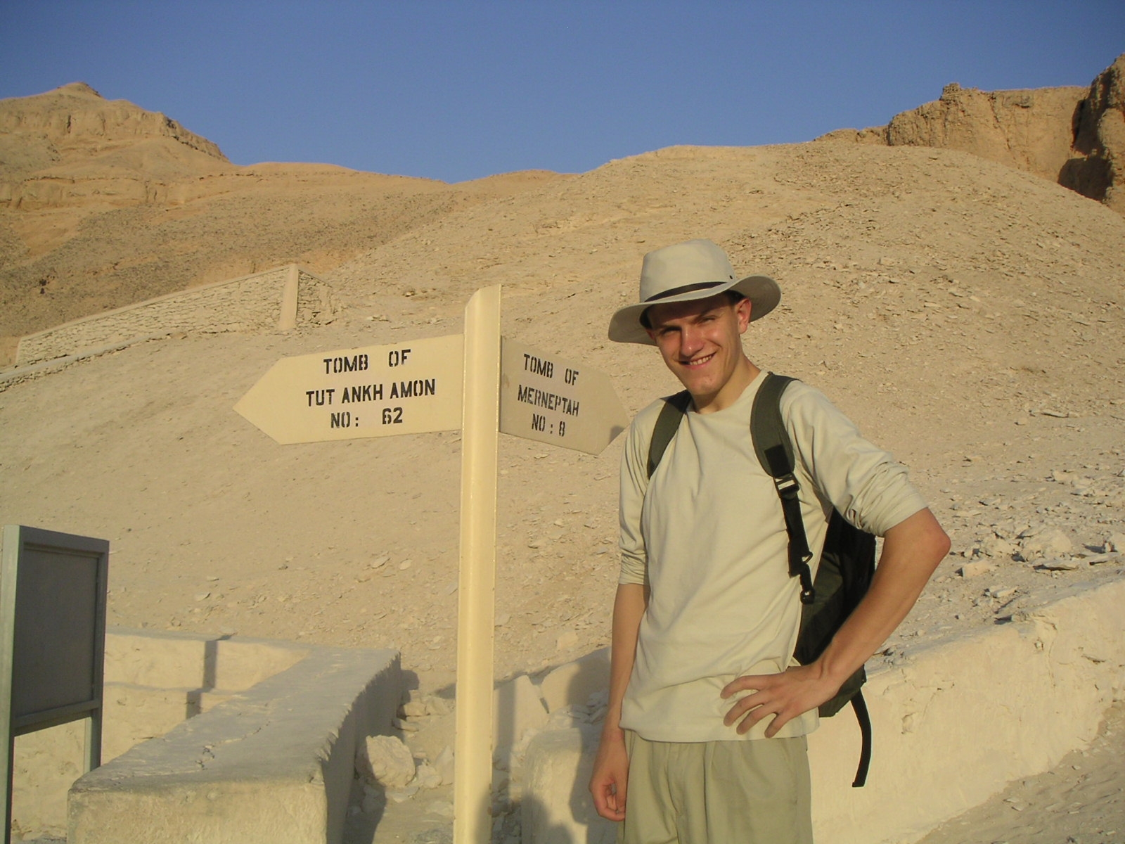 A picture of me, smiling, wearing a light-coloured wide-brimmed sun hat, long-sleeved shirt, and backpack stands beside a signpost in a sunlit desert landscape of pale limestone hills, in Egypt's Valley of the Kings. One hand rests on my hip as I pose for the camera. The signpost points in two directions and reads: "TOMB OF TUT ANKH AMON NO: 62" on the left arrow and "TOMB OF MERNEPTAH NO: 8" on the right arrow. Low white stone walls border the path in the foreground, while the barren rocky hills rise behind under a cloudless deep blue sky. Warm late-afternoon sunlight casts long shadows across the scene.