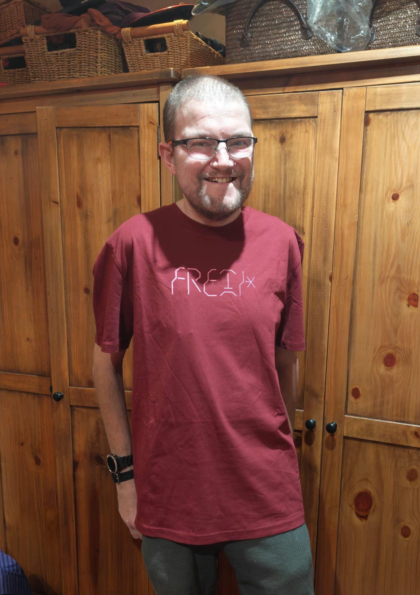 A picture of me smiling at the camera in front of wooden cupboards, wearing glasses and a burgundy Fluke T-shirt with the word “Freak” across the chest in a heavily stylised pale-pink font.
