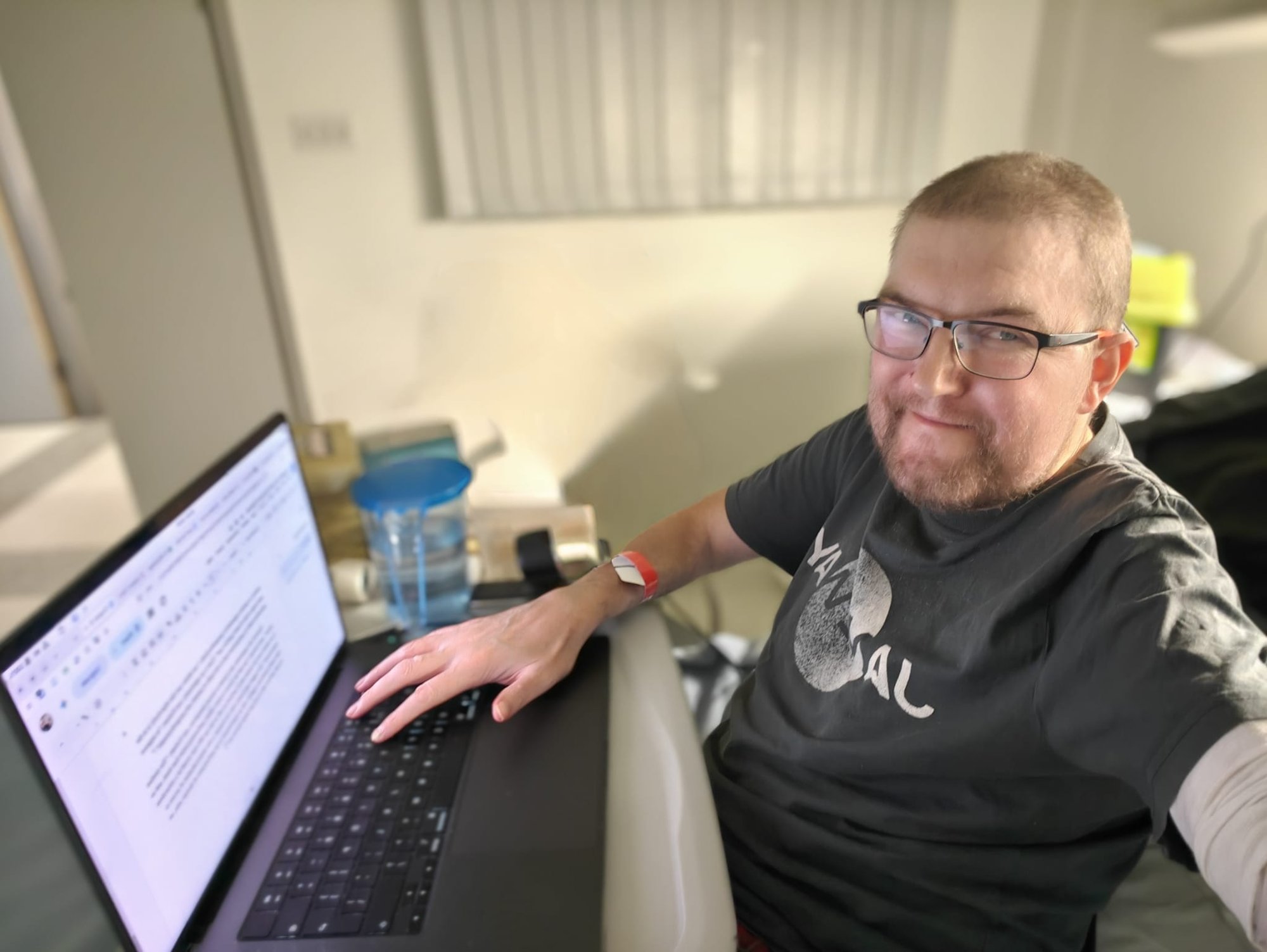 A picture of me, Martin Paul Eve, writing at a computer, in a hospital, with a hospital wrist band and an Apple Mac computer running a Linux virtual machine. I am a white man, here with germinative beard and longer hair than usual by a long way (but still fairly short). I am wearing a Janeway t-shirt.